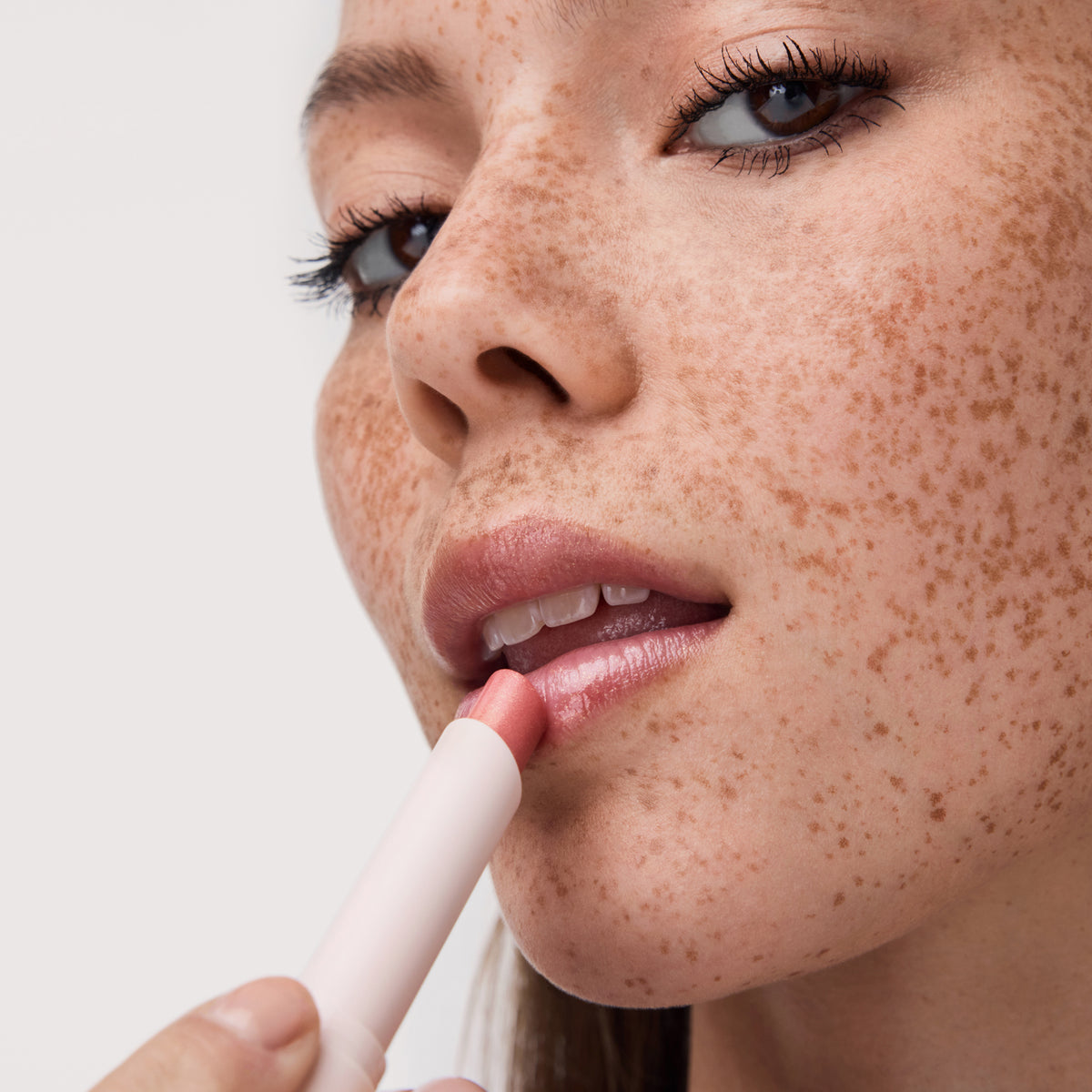 Eine sommersprossige Frau trägt den Diamond Glaze Gloss Stick von Catrice Cosmetics auf ihre Lippen auf, der in einer Nahaufnahme ihres Gesichts zu sehen ist.