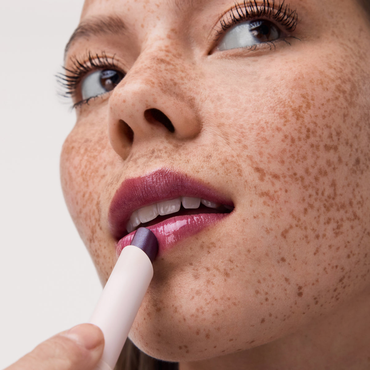 Nahaufnahme einer Frau mit Sommersprossen, die einen schimmernden burgunderfarbenen Diamond Glaze Gloss Stick auf ihre Lippen aufträgt.