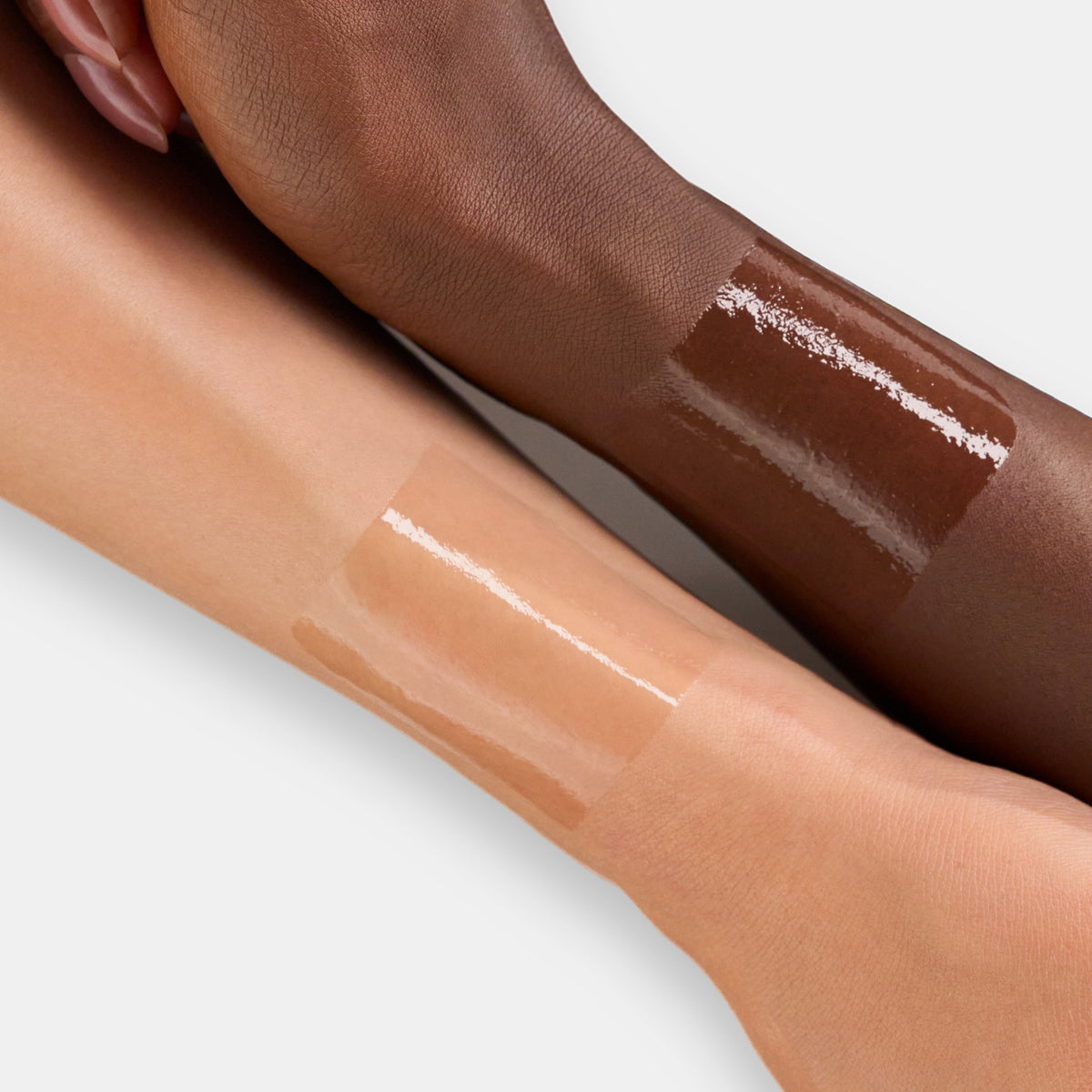 Zwei Unterarme mit unterschiedlichen Hauttönen, beide mit einem aufgetragenen, glänzenden Swatch einer Lippenmaske oder eines Glosses.