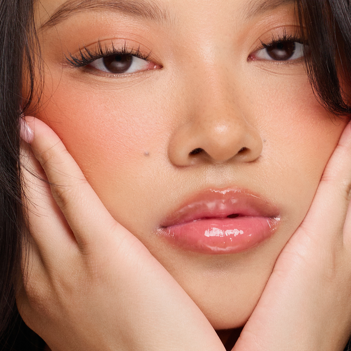 Nahaufnahme einer Frau mit glänzenden Lippen unter Verwendung von Dominique Cosmetics Juicy Lip Balm, pfirsichfarbenem Rouge, Eyeliner und Händen, die ihr Gesicht umrahmen.