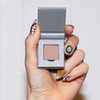Eine Hand mit kunstvollen Nägeln hält The Single von LH Cosmetics, eine offene graue Dose mit nudefarbenem Puder darin.