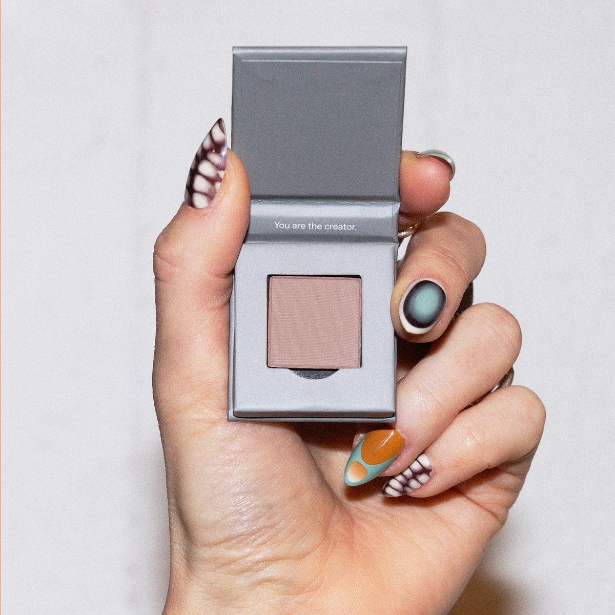 Eine Hand mit kunstvollen Nägeln hält The Single von LH Cosmetics, eine offene graue Dose mit nudefarbenem Puder darin.