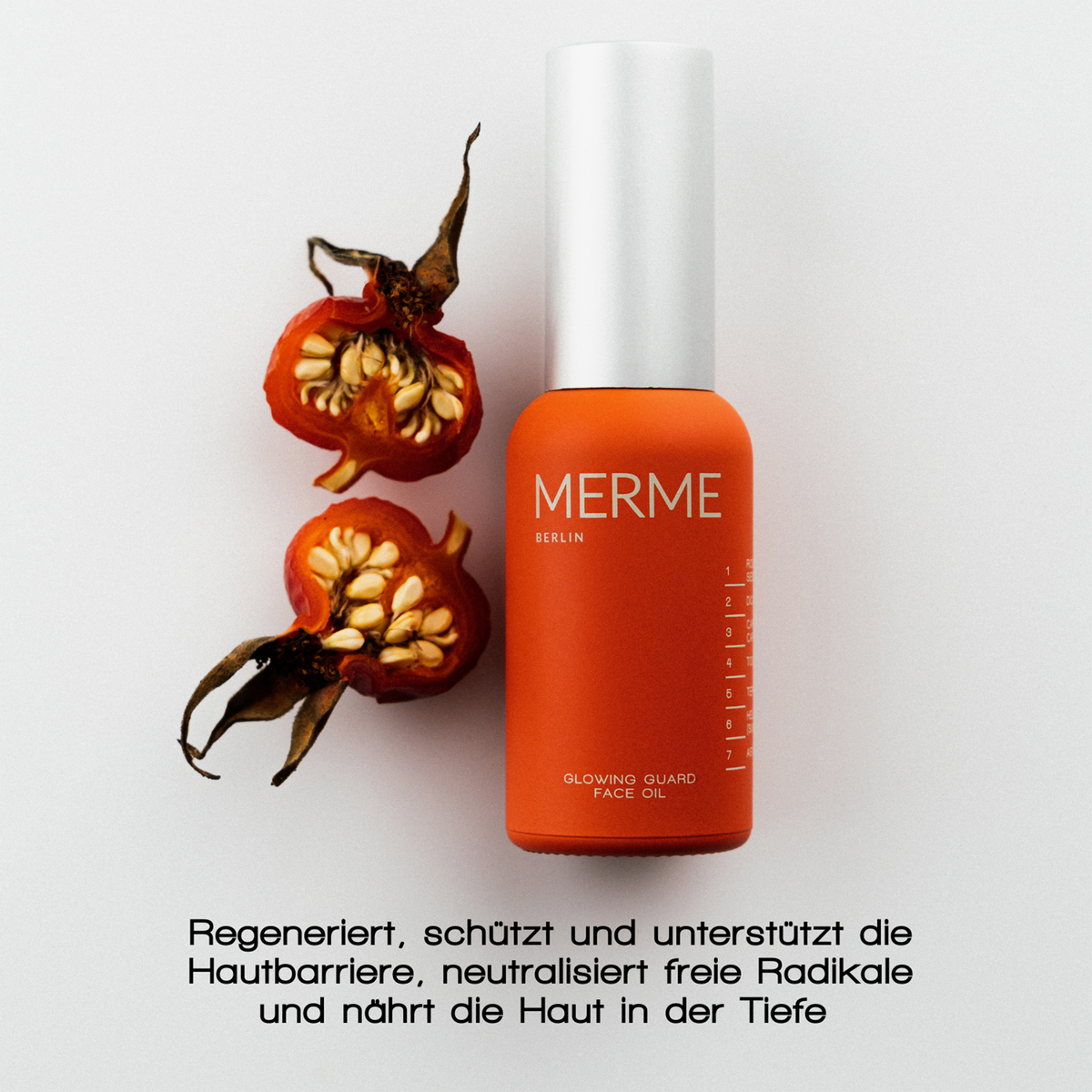 Eine Flasche MERME Glowing Guard Face Oil steht neben zwei halbierten Hagebutten auf einem weißen Hintergrund.