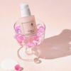Eine Flasche numbuzin No.4 Collagen 73% Pudding Serum befindet sich in einem Glasbecher mit rosa und klaren Perlen auf einem hellrosa Hintergrund.