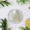 Das No.5 Goodbye Blemish Serum von numbuzin befindet sich auf einer Marmorplatte, umgeben von grünen Blättern und einem Glas Orangensaft, alles auf weißem Stoff.