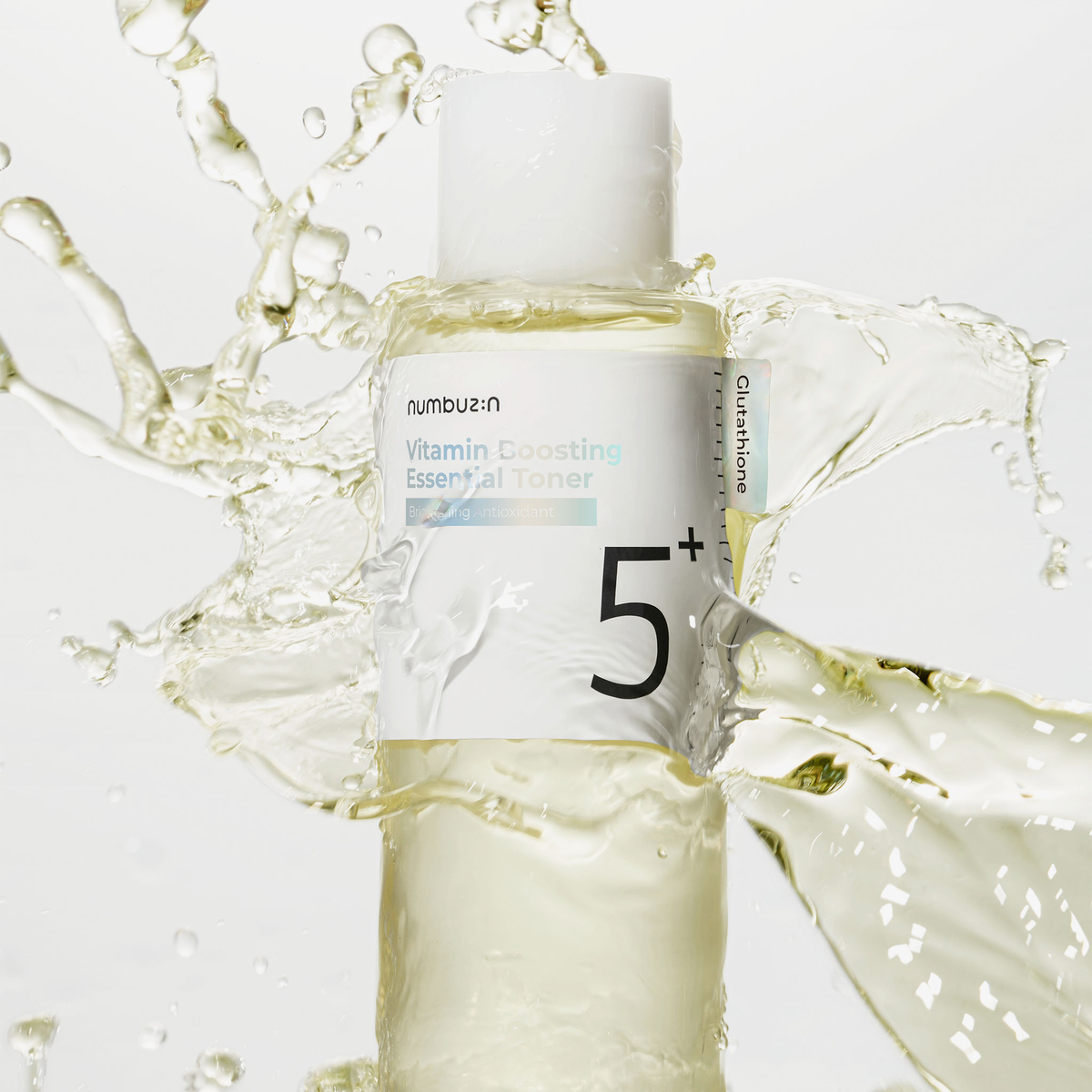 Eine durchsichtige Flasche des numbuzin No.5+ Vitamin Boosting Essence Toner ist vor einem weißen Hintergrund mit Flüssigkeit bespritzt.