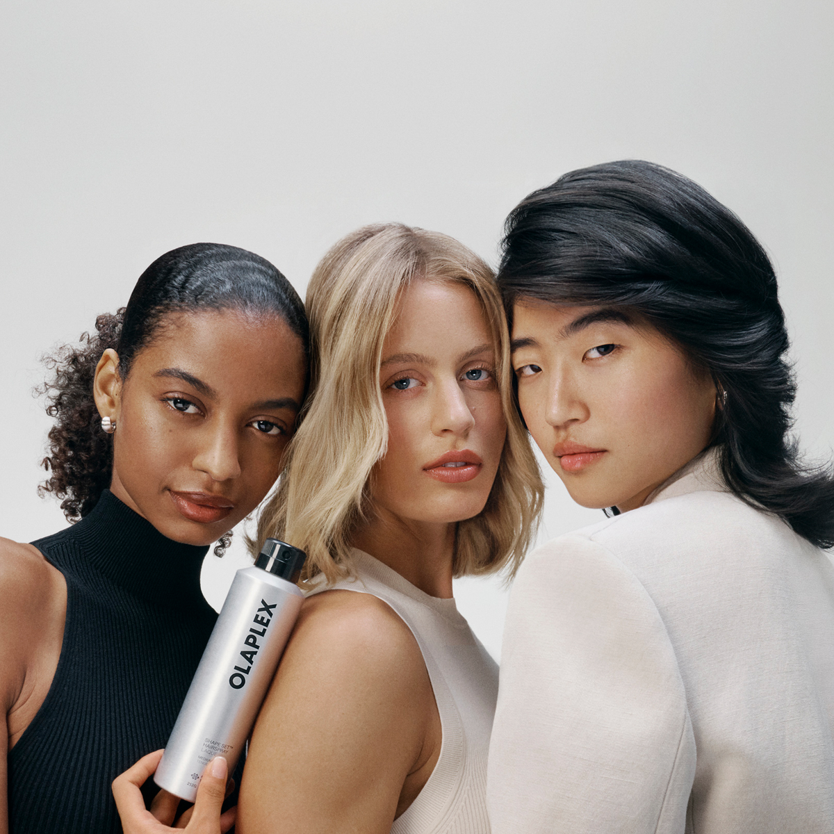 Drei Frauen posieren gemeinsam, eine von ihnen hält eine hohe silberne Flasche OLAPLEX Shape Set Hairspray in der Hand.