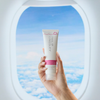 Eine Hand hält eine Philip-Kingsley-Badekappe vor einem Flugzeugfenster, hinter dem sich ein blauer Himmel und Wolken zeigen.