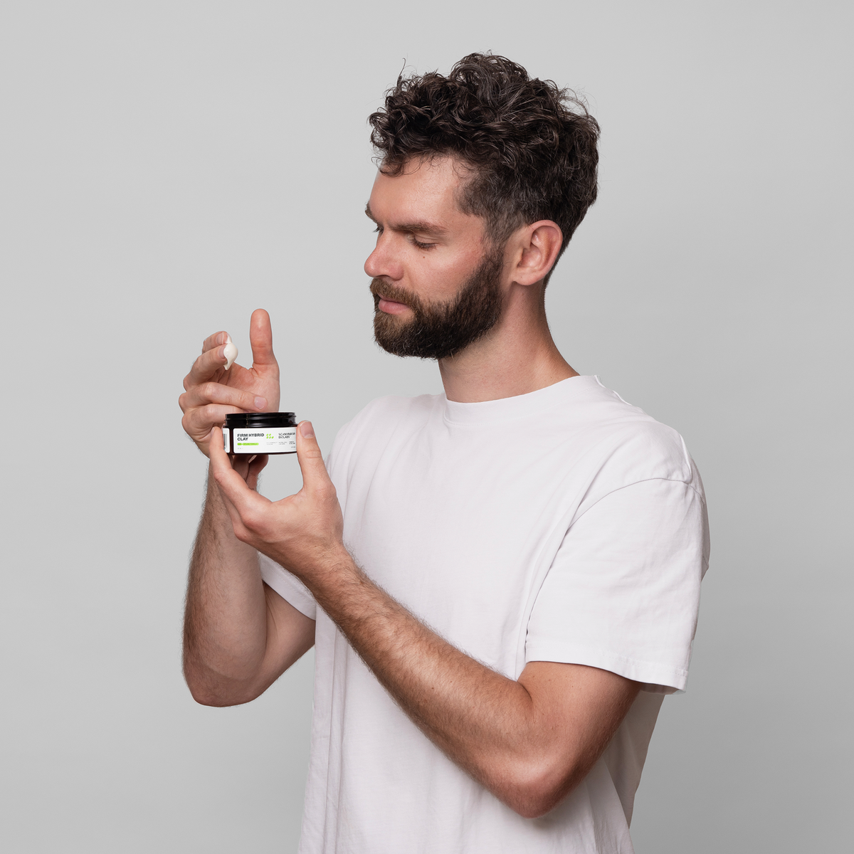 Ein Mann in einem weißen T-Shirt trägt vor einem neutralen grauen Hintergrund Scandinavian Biolabs Firm Hybrid Clay aus einem Tiegel auf.