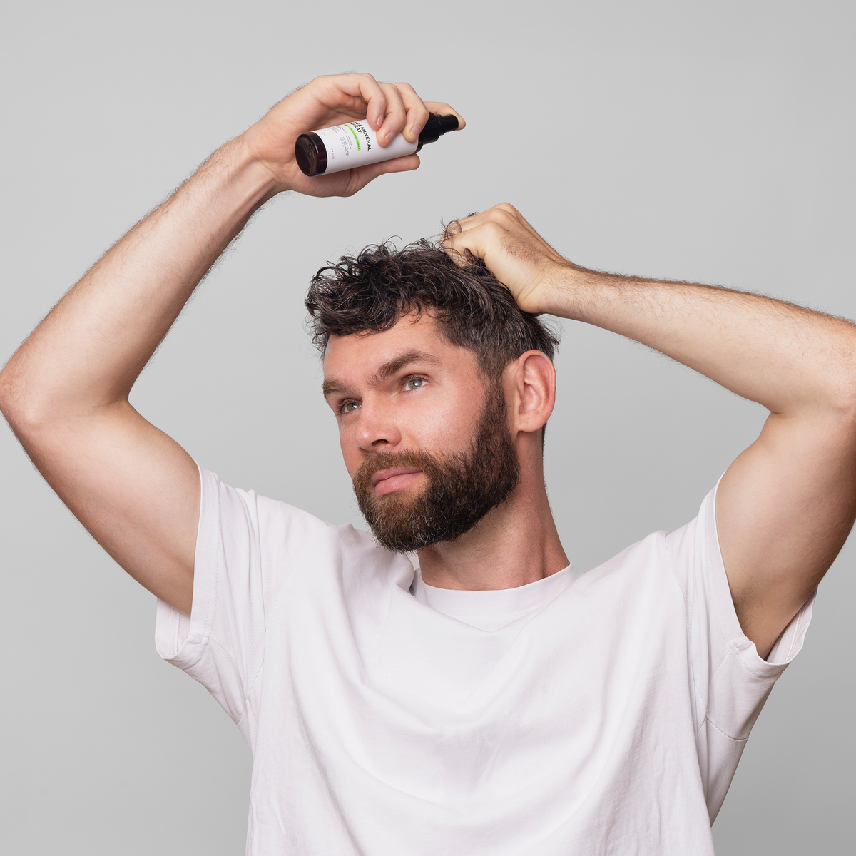 Ein Mann in einem weißen Hemd trägt Scandinavian Biolabs Sea Mineral Spray aus einer Sprühflasche auf sein lockiges Haar auf, vor einem grauen Hintergrund.