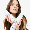 Eine Frau mit langen braunen Haaren hält zwei Flaschen SHYNE Cloud Shampoo vor einem weißen Hintergrund.