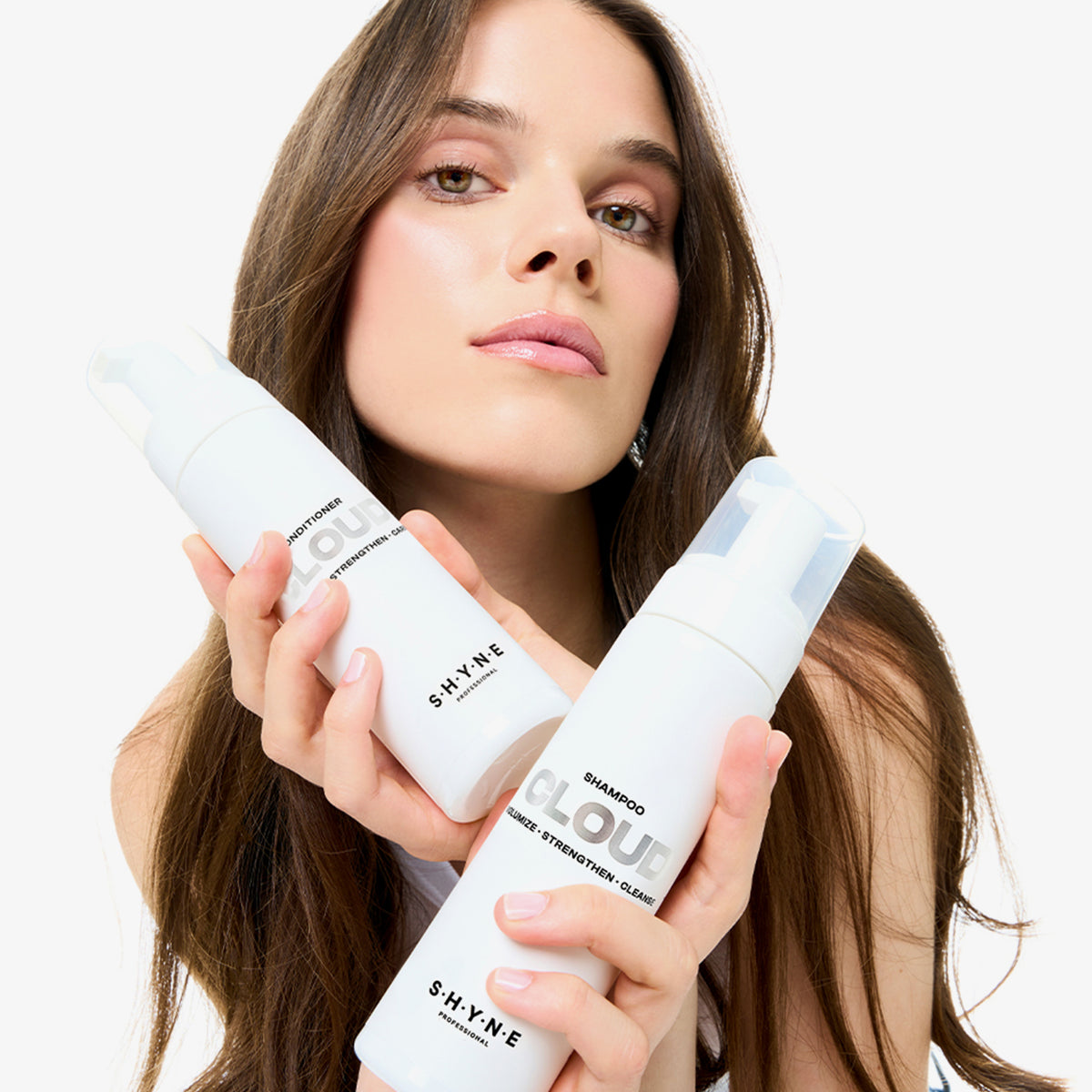 Eine Frau mit langen braunen Haaren hält zwei Flaschen SHYNE Cloud Shampoo vor einem weißen Hintergrund.