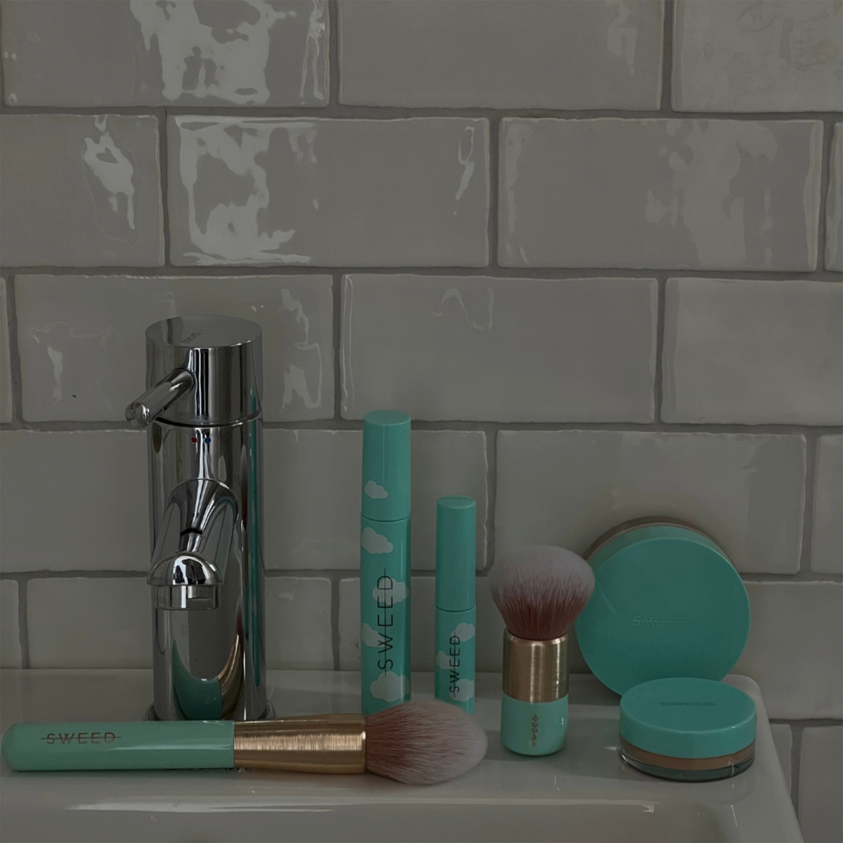 Ein mintgrüner Foundation-Pinsel von SWEED liegt auf einem Waschbecken vor einem Hintergrund aus weißen Metrofliesen, daneben liegen passende Make-up-Produkte.