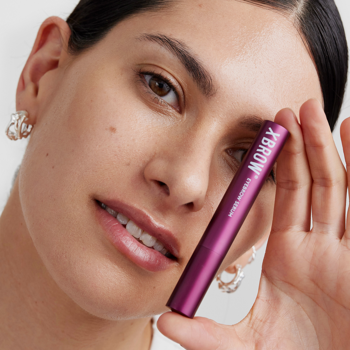 Eine Frau hält eine violette Tube XLASH Xbrow Augenbrauenserum neben ihr Gesicht und lächelt sanft in die Kamera.