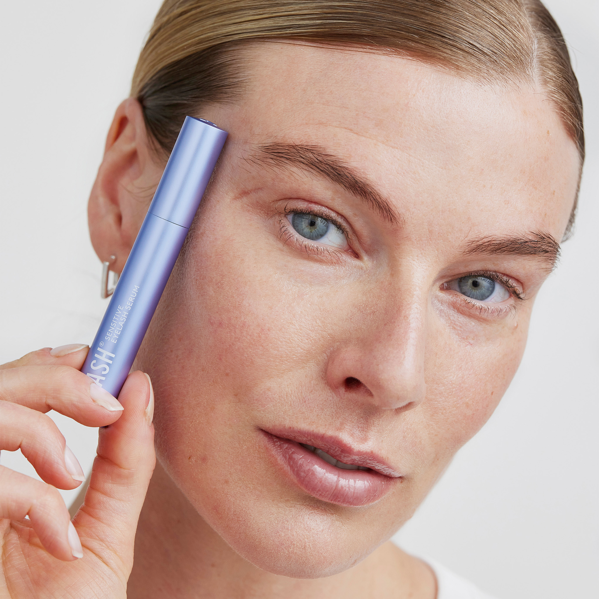 Eine Person hält eine Tube „Xlash Sensitive Eyelash Serum“ von XLASH nahe an ihre Augenbraue und blickt in die Kamera.