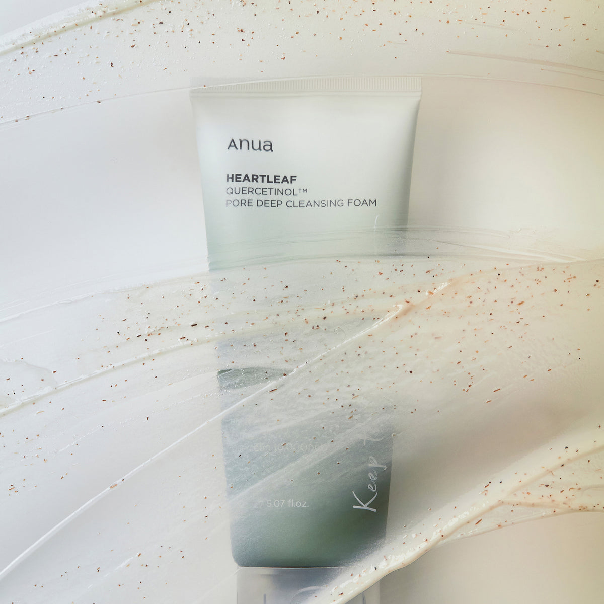 Eine Tube Heartleaf Quercetinol Pore Deep Cleansing Foam liegt auf einer cremigen, strukturierten Oberfläche mit sichtbaren Peelingpartikeln.