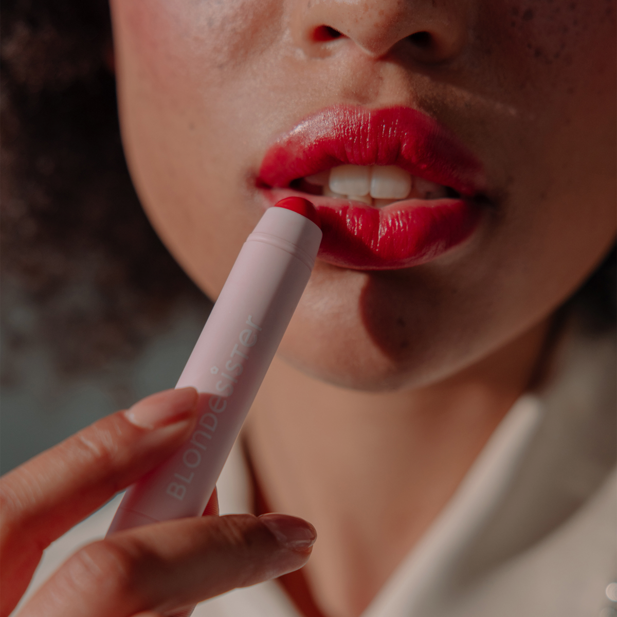 Jemand verwendet „BLONDESISTER 2 IN 1 – IT’S UP TO YOU LIP OR CHEEK“ und trägt es aus einer hellrosa Tube auf die Lippen auf.