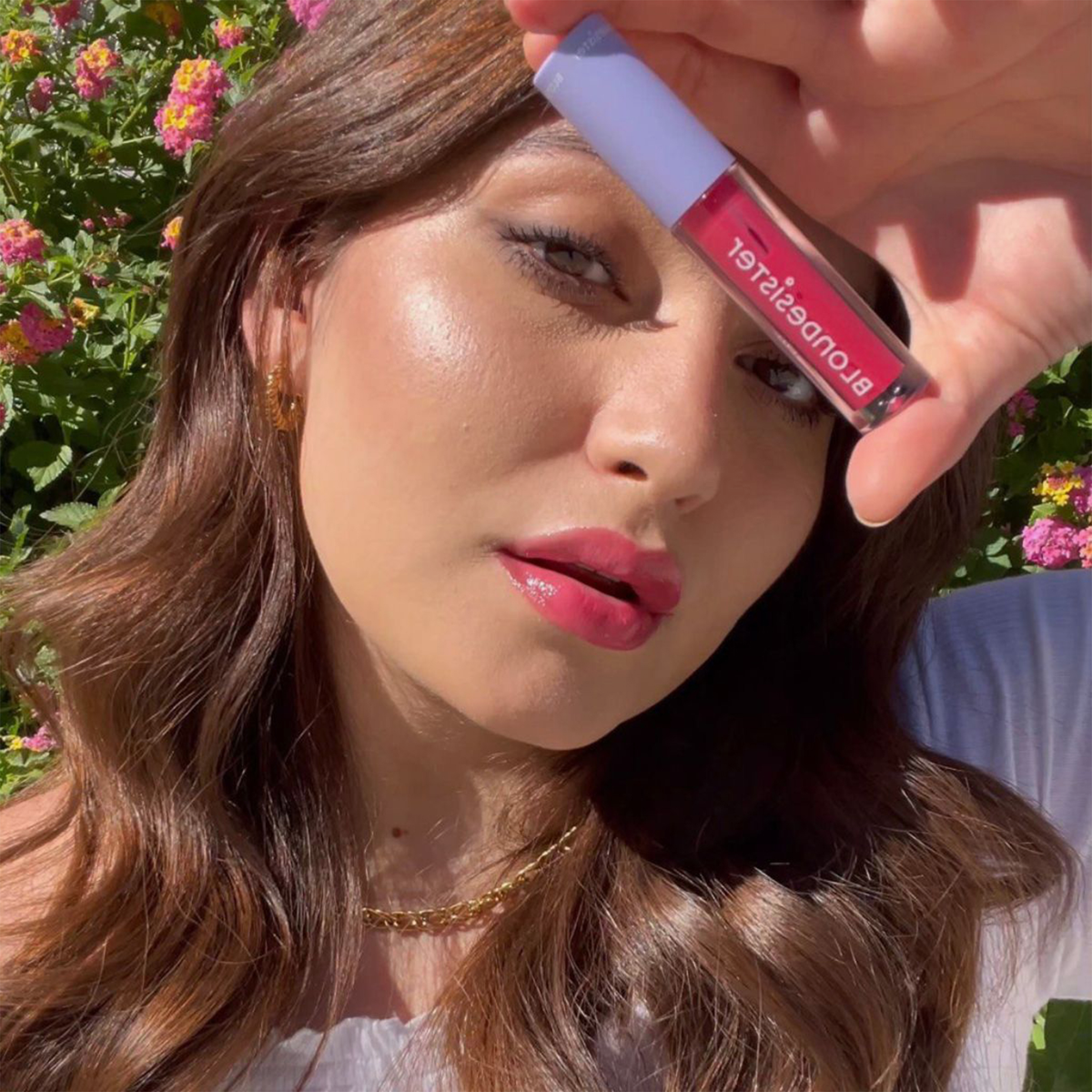 Eine Frau mit welligem braunem Haar trägt das „BLONDESISTER 2 IN 1 SWEET GLOSS TINTED LIP OIL“ von „flowering bushes“ auf – ihre Lippen schimmern in einem zarten Rosa.