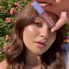 Eine Frau hält das „BLONDESISTER 2 IN 1 SWEET GLOSS TINTED LIP OIL“ nahe an ihr Gesicht, im Freien, hinter ihr sind rosa und gelbe Blumen zu sehen.