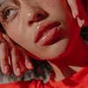 Eine Nahaufnahme von glänzenden Lippen mit Sommersprossen in rotem Licht, dazu ein rotes Oberteil und das „BLONDESISTER 3 IN 1 CUTE LIPPY LIP OIL“.