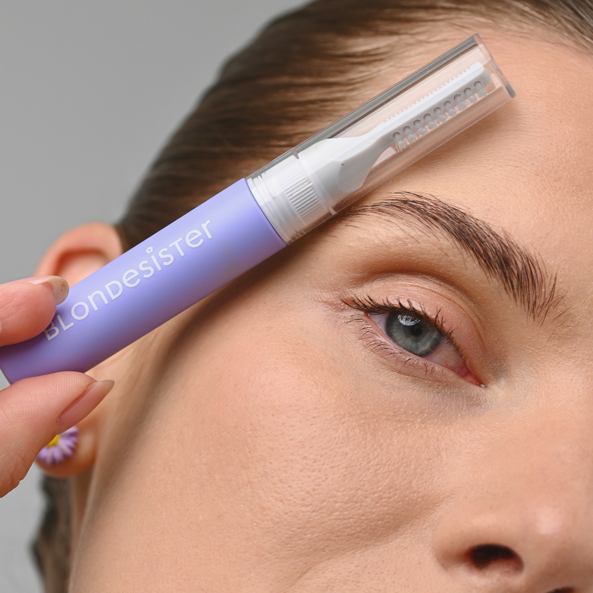 Eine Frau hält das „3 IN 1 FIX&GO BROW GEL“ von BLONDESISTER in der Nähe ihrer Augenbraue.
