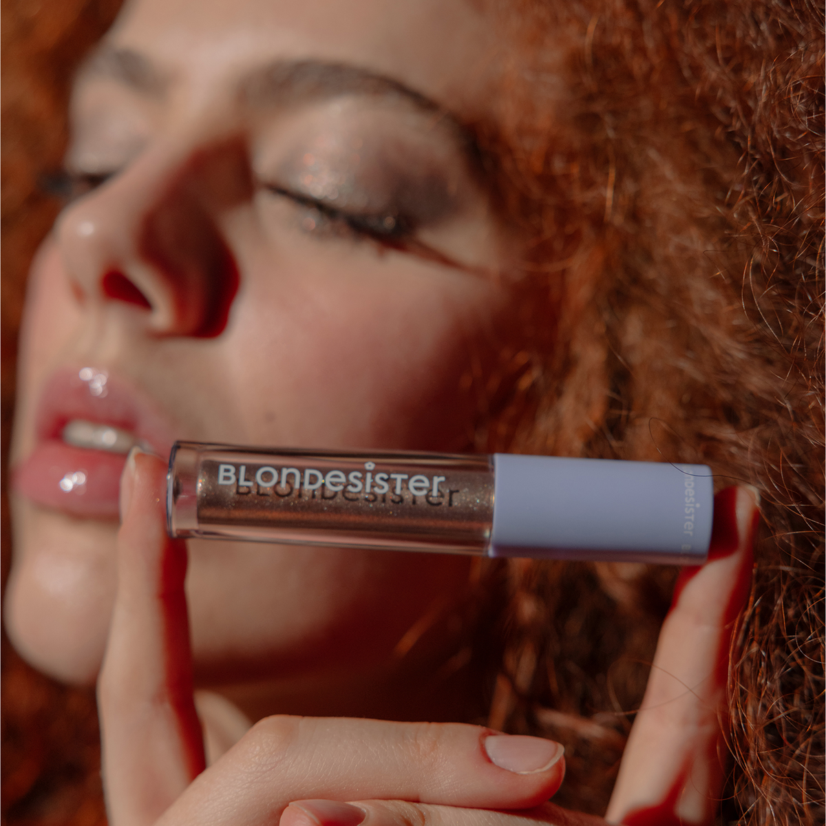 Eine Frau mit lockigem Haar hält einen „BLONDESISTER GLOWEYE EYESHADOW“ nahe an ihre Lippen, die Augen sind geschlossen.