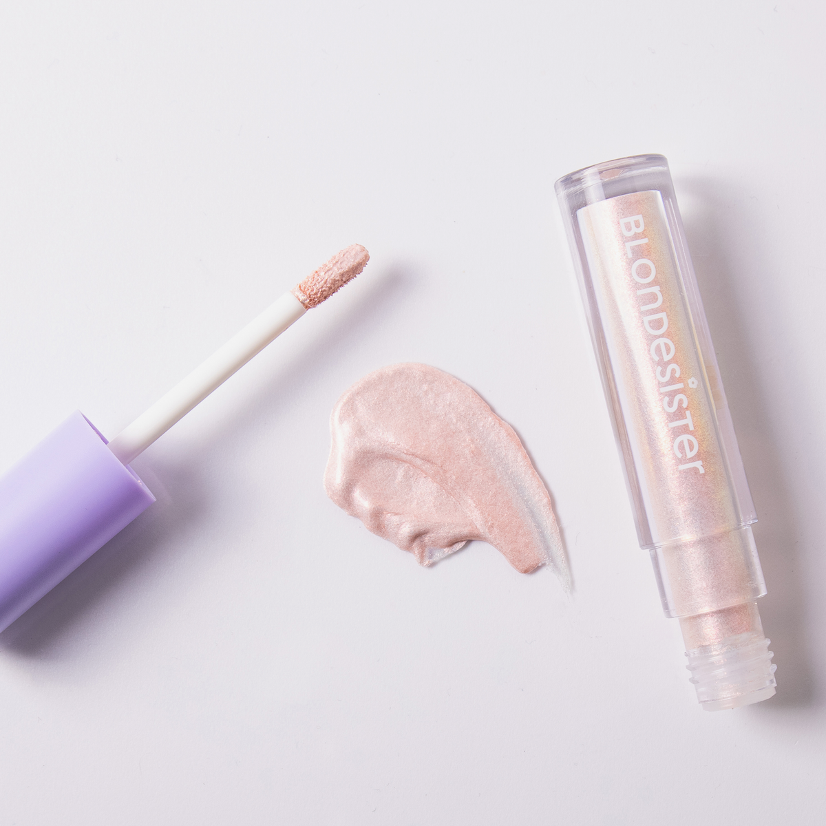 Eine Tube „BLONDESISTER GLOWEYE EYESHADOW“ mit violettem Verschluss liegt neben einem Farbmuster auf einer weißen Unterlage.