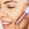 Eine lächelnde Person, deren Augen mit schimmerndem Lidschatten geschminkt sind, hält eine Tube „GLOWEYE EYESHADOW“ von BLONDESISTER nahe an ihr Gesicht.