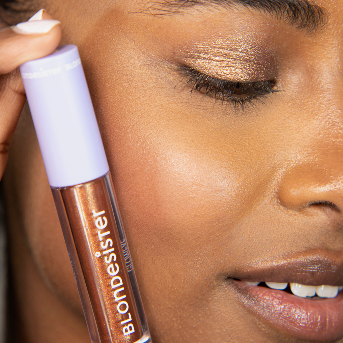 Eine Nahaufnahme einer Person, die eine Tube „BLONDESISTER GLOWEYE EYESHADOW“ neben ihrem metallisch-goldenen Lidschatten-Look hält.