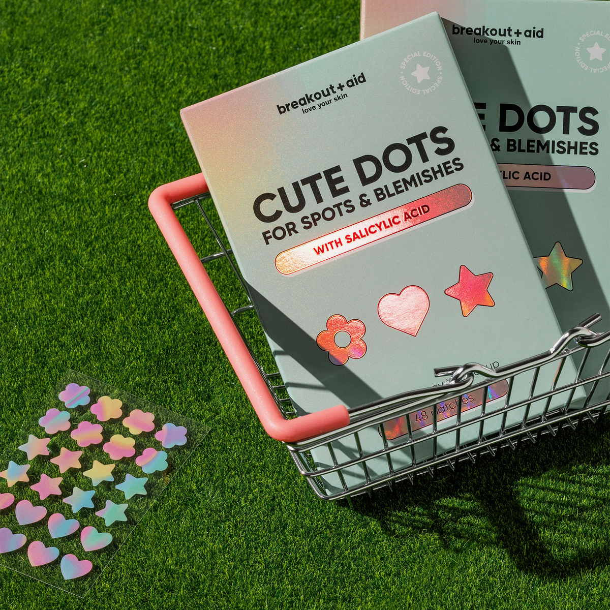 Zwei Schachteln Cute Dots gegen Hautunreinheiten und Pickel sowie ein Bogen mit stern- und blumenförmigen Aufklebern in einem kleinen Einkaufskorb aus Metall auf dem Rasen.