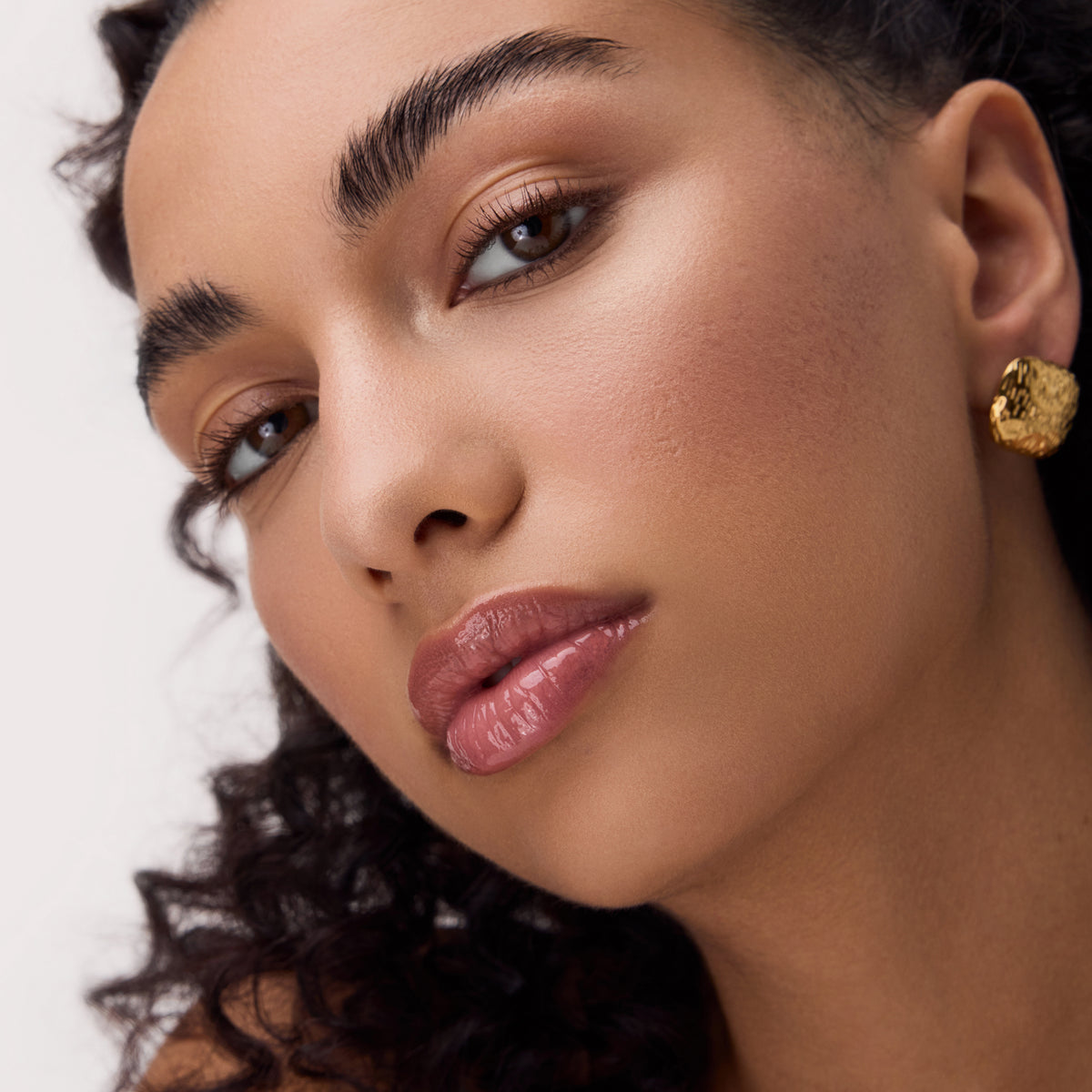 Eine Frau mit lockigem Haar, natürlichem Make-up und goldfarbenen Ohrringen trägt das Catrice Cosmetics Colour Cushion Juicy Lip Oil und blickt in die Kamera.