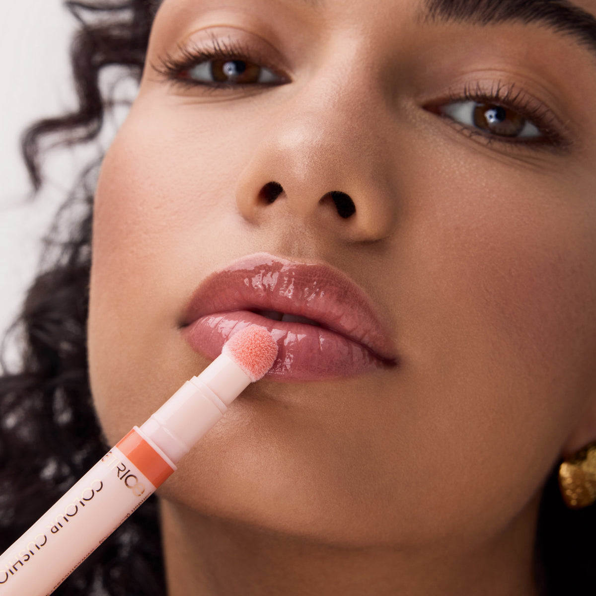 Nahaufnahme einer Frau, die Catrice Cosmetics Colour Cushion Juicy Lip Oil, ein rosafarbenes, glänzendes Lippenprodukt, mit einem Applikator auf ihre Lippen aufträgt.