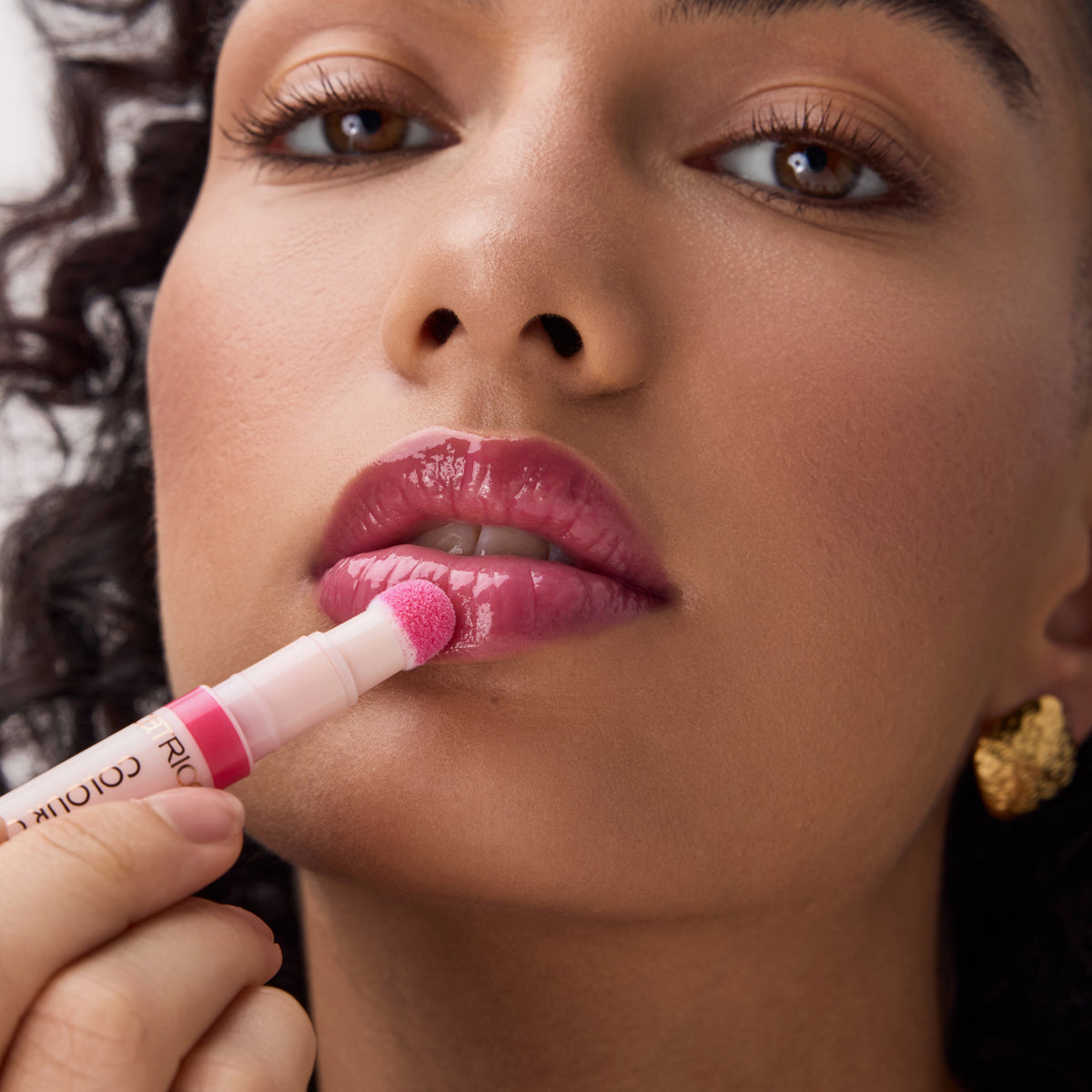 Frau trägt mit einem Applikator ein glänzendes pinkes Lip Oil auf ihre Lippen auf, Nahaufnahme mit Fokus auf Lippen und Produkt.