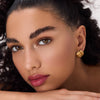 Porträt einer Frau mit glänzendem, kirschrotem Lippenöl, sanftem Make-up und goldenen Ohrringen, aufgetragen mit dem Catrice Colour Cushion Lip Oil.