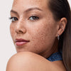 Eine Frau mit Sommersprossen, braunen Augen und Ohrringen posiert vor einem hellen Hintergrund und trägt den Catrice Cosmetics Diamond Glaze Gloss Stick.