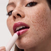 Nahaufnahme einer Frau mit Sommersprossen, die einen schimmernden pinken Diamond Glaze Gloss Stick von Catrice auf ihre Lippen aufträgt.