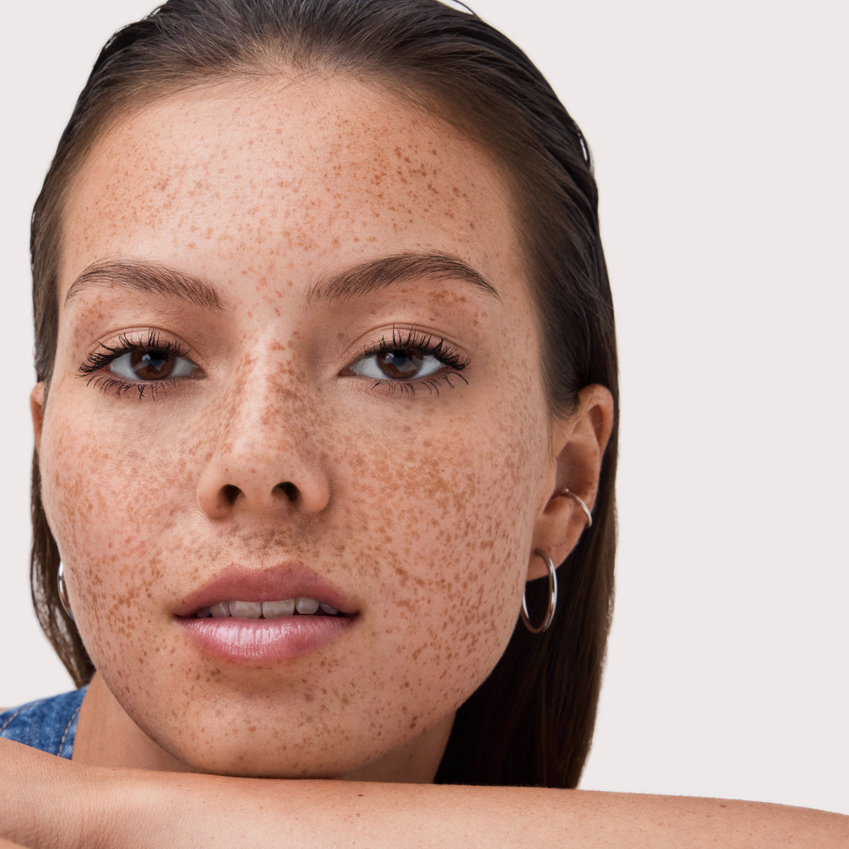 Porträt einer Frau mit vielen Sommersprossen, dezent glänzenden Lippen und dezentem Make-up, sie stützt ihr Kinn auf den Arm und blickt ruhig in die Kamera.
