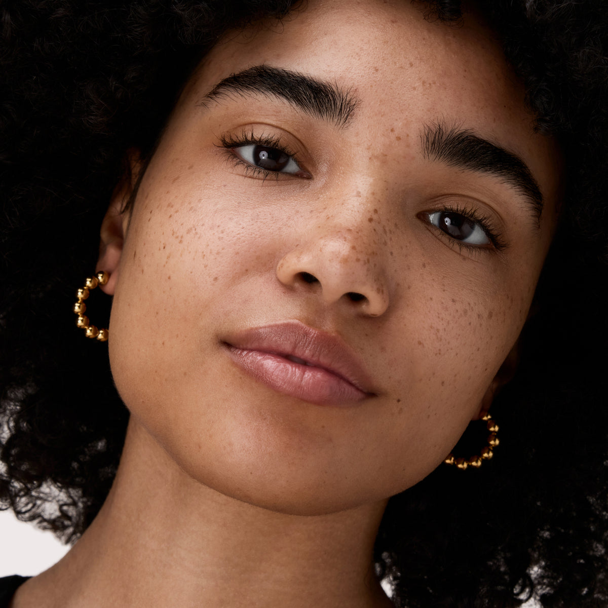 Eine selbstbewusste Person mit lockigem Haar, Sommersprossen und goldenen Ohrringen modelliert das Under Eye Brightener Liquid von Catrice Cosmetics.