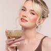 Eine Frau mit taufrischem Make-up lächelt in die Kamera, in der Hand ein Getränk und Dominique Cosmetics Juicy Lip Balm.