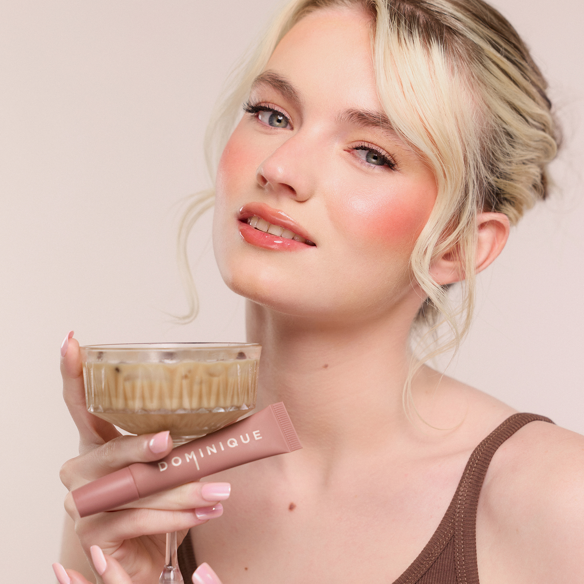 Eine Frau mit taufrischem Make-up lächelt in die Kamera, in der Hand ein Getränk und Dominique Cosmetics Juicy Lip Balm.