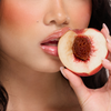 Eine Person mit glänzenden Lippen vom Juicy Lip Balm von Dominique Cosmetics hält einen angebissenen Pfirsich an den Mund, so dass der Kern sichtbar wird.