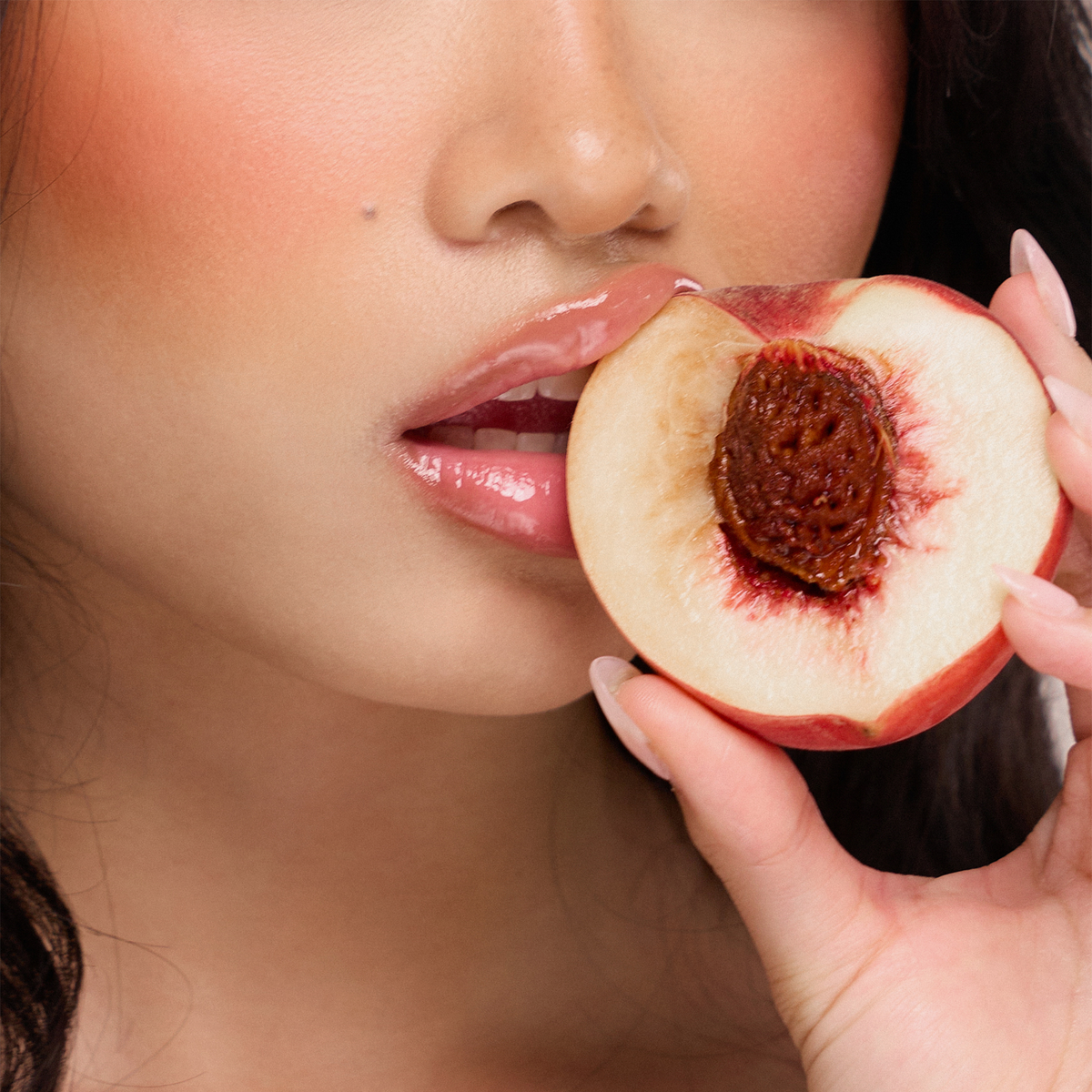 Eine Person mit glänzenden Lippen vom Juicy Lip Balm von Dominique Cosmetics hält einen angebissenen Pfirsich an den Mund, so dass der Kern sichtbar wird.
