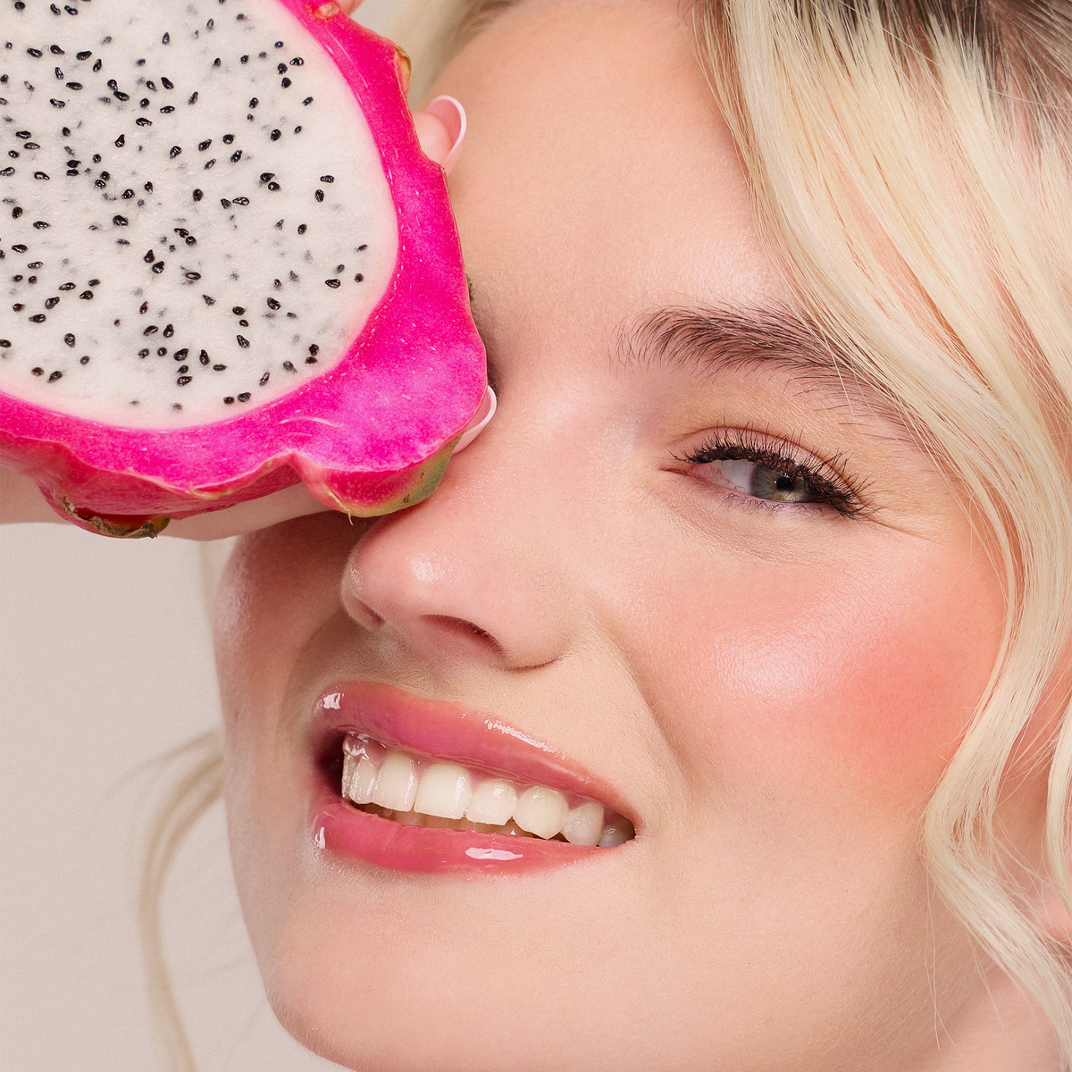 Lächelnde Frau hält Juicy Lip Balm von Dominique Cosmetics in der Nähe ihres Auges vor einem hellen Hintergrund.