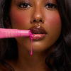 Eine Frau mit glänzenden Lippen trägt Dominique Cosmetics Juicy Lip Balm aus einer rosafarbenen Tube auf, eine leuchtend pinke Flüssigkeit auf ihren Lippen.