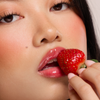 Eine Frau mit glänzenden Lippen von Dominique Cosmetics Juicy Lip Balm hält eine reife Erdbeere an ihren Mund.