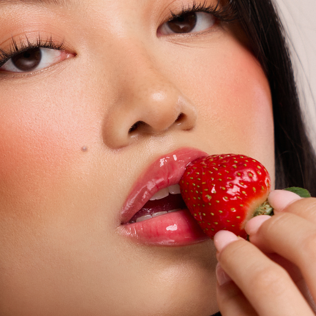 Eine Frau mit glänzenden Lippen von Dominique Cosmetics Juicy Lip Balm hält eine reife Erdbeere an ihren Mund.