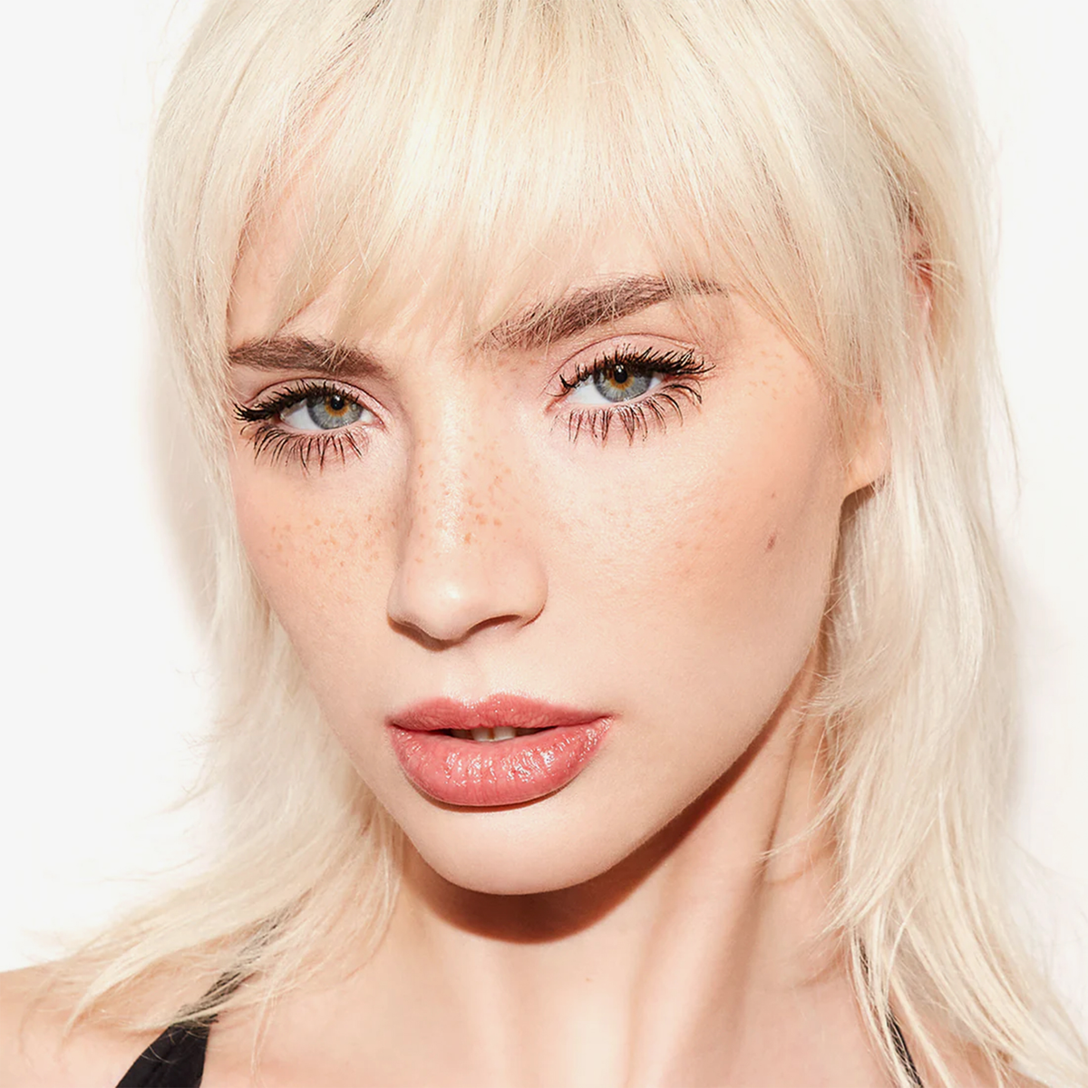 Nahaufnahme einer Frau mit platinblondem Haar und Sommersprossen, die Freck OG von Freck Beauty trägt, vor einem weißen Hintergrund.