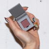 Eine Hand mit Nailart hält eine geöffnete LH Cosmetics The Single Puderdose mit mauvefarbenem Puder vor einem weißen Hintergrund.