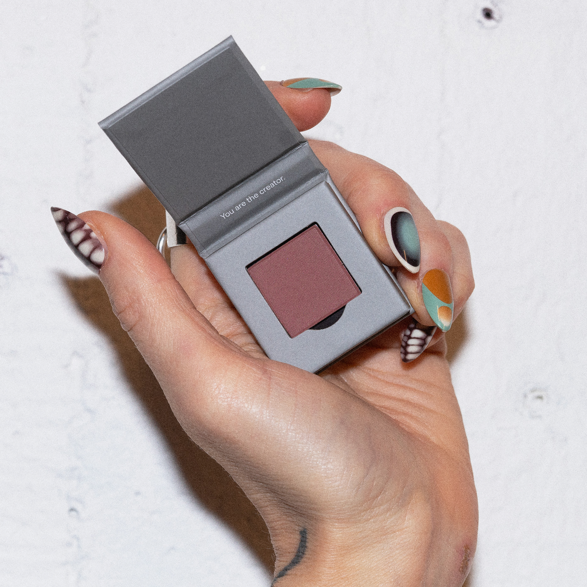 Eine Hand mit Nailart hält eine geöffnete LH Cosmetics The Single Puderdose mit mauvefarbenem Puder vor einem weißen Hintergrund.