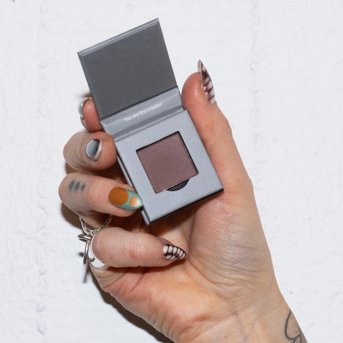 Eine Hand mit Nailart hält eine geöffnete LH Cosmetics The Single mauve eyeshadow compact vor einem weißen Hintergrund.